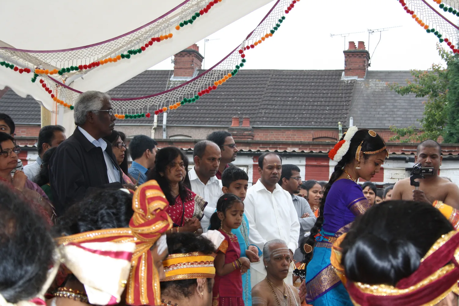 The procession in the street outside, kalasam pots carried past the brick houses.