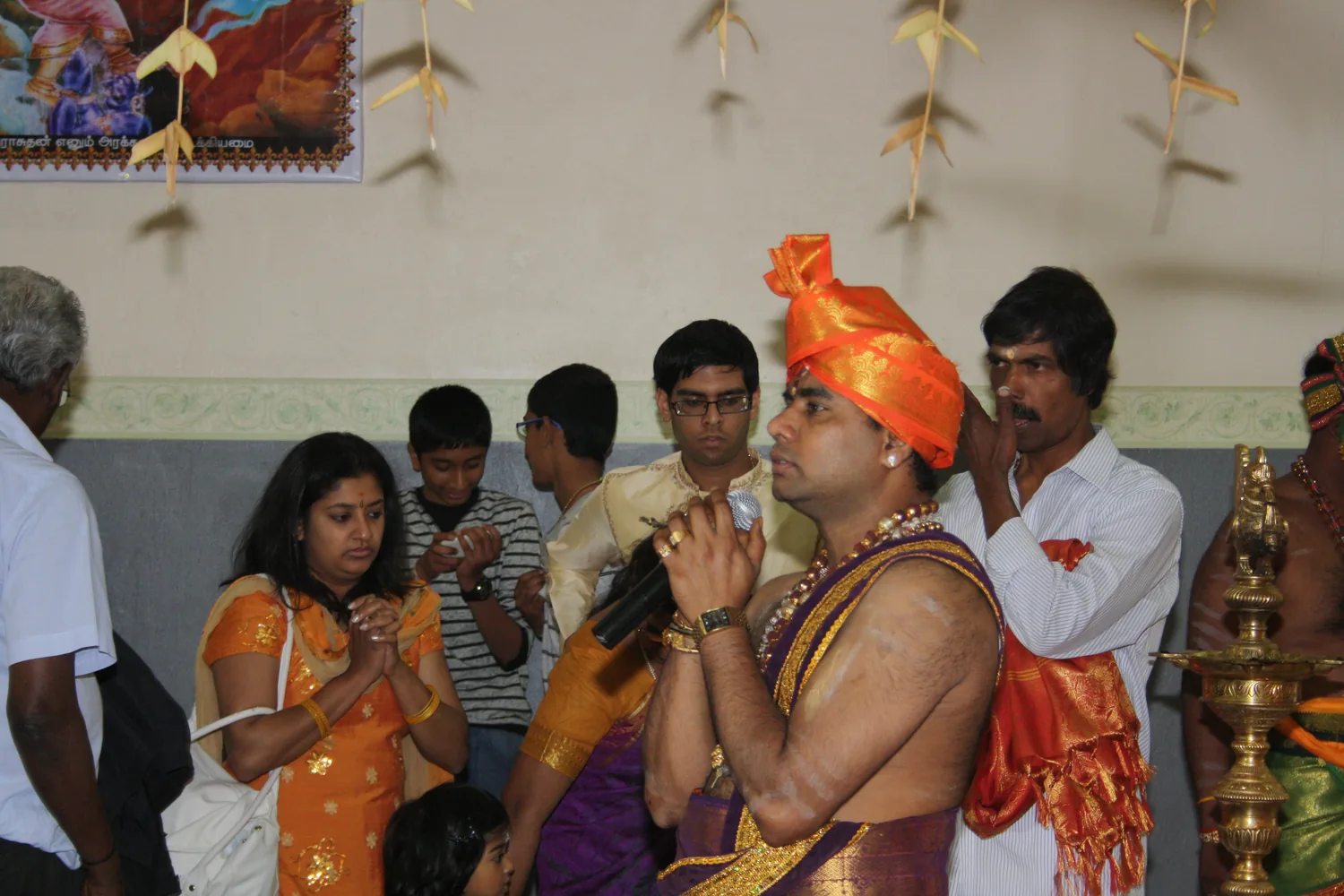 A priest with palms joined, the hall behind him.